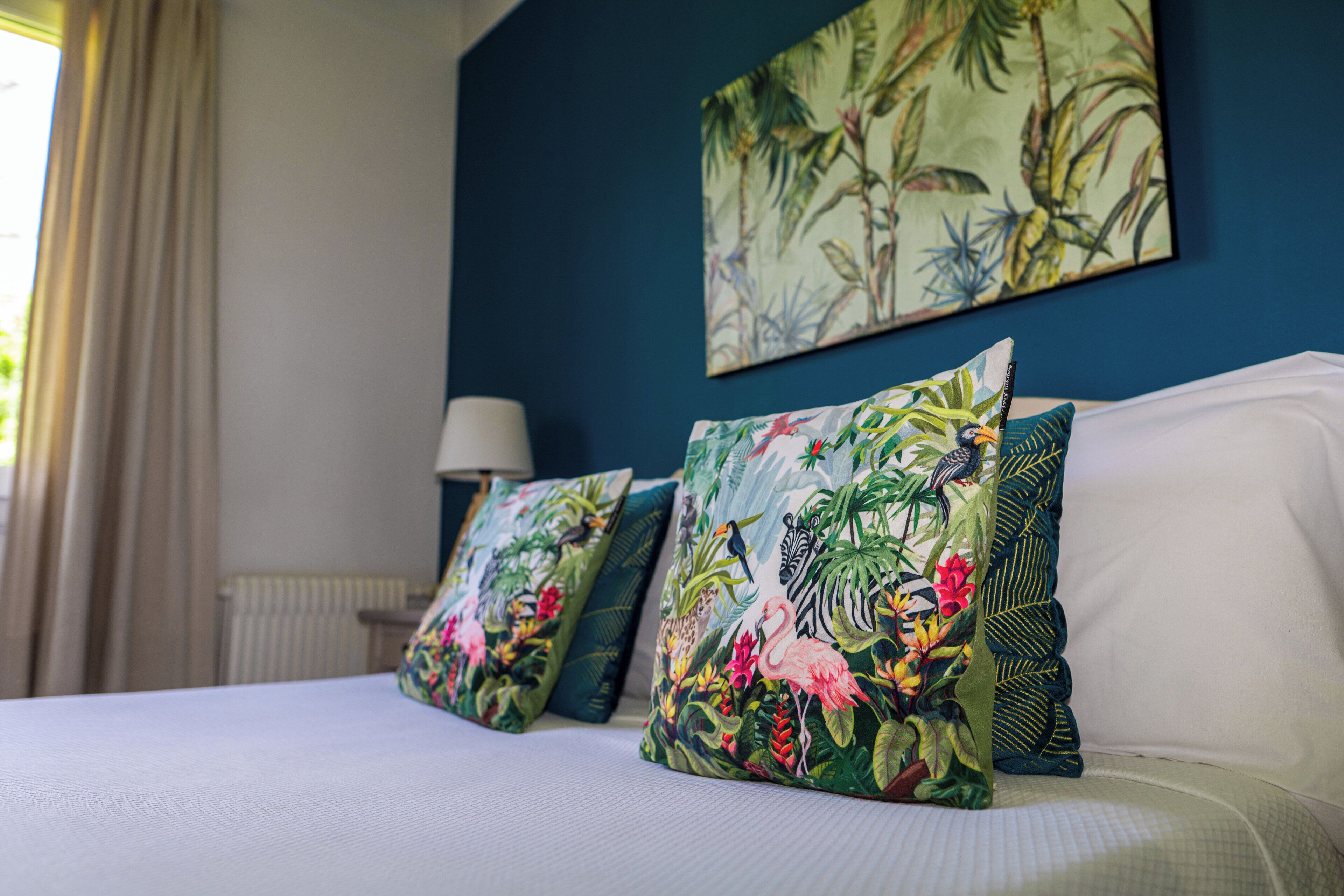 A bed with floral-patterned pillows, a nightstand with a lamp, and a tropical-themed painting on the wall.
