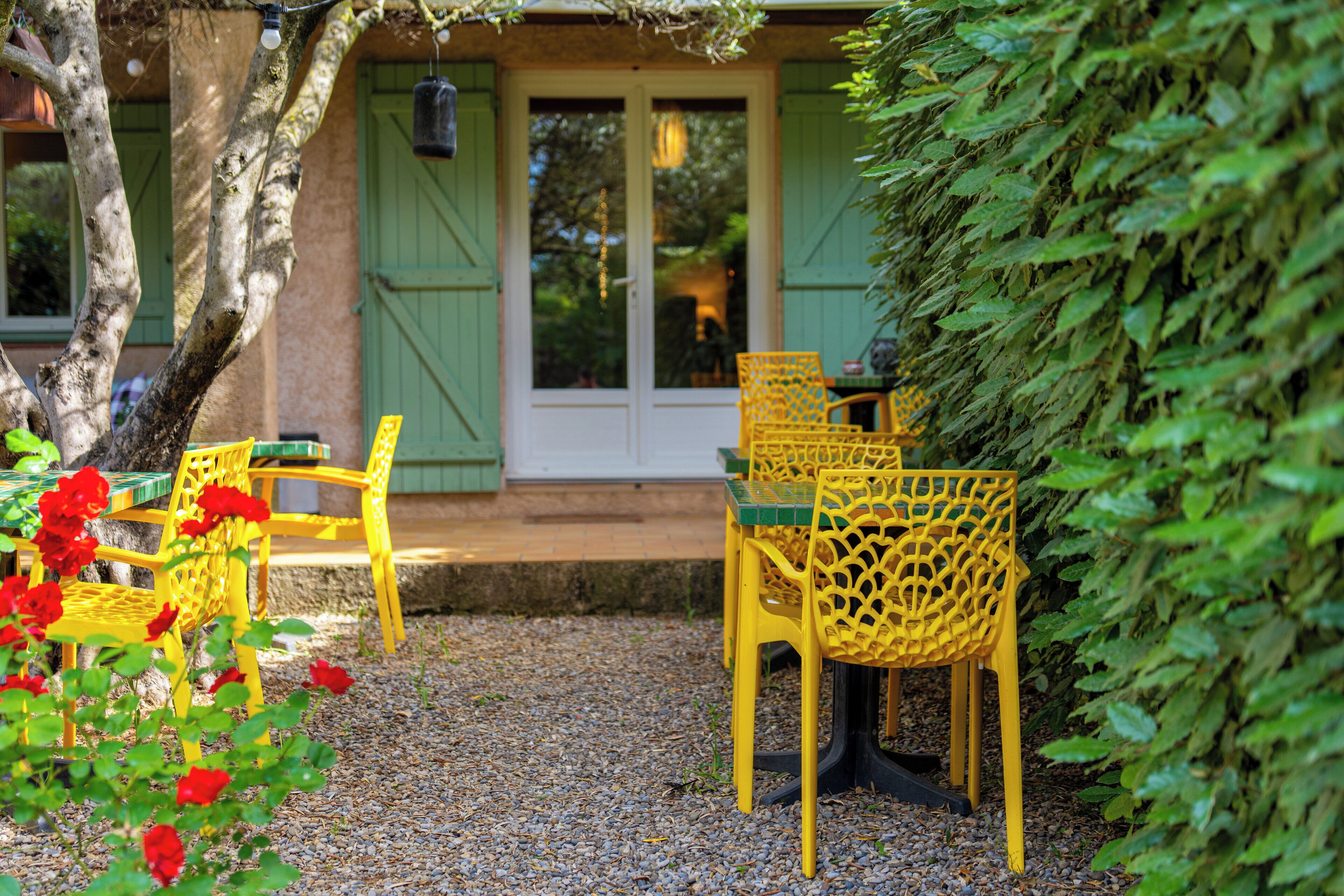 An outdoor seating area with yellow chairs and a table, surrounded by greenery and a gravel path.