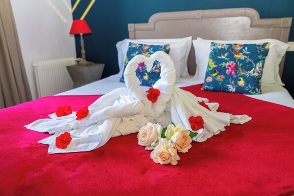Una cama con una colcha roja, almohadas blancas y una almohada estampada con motivos florales. Hay toallas blancas dispuestas en forma de cisne con flores rojas.