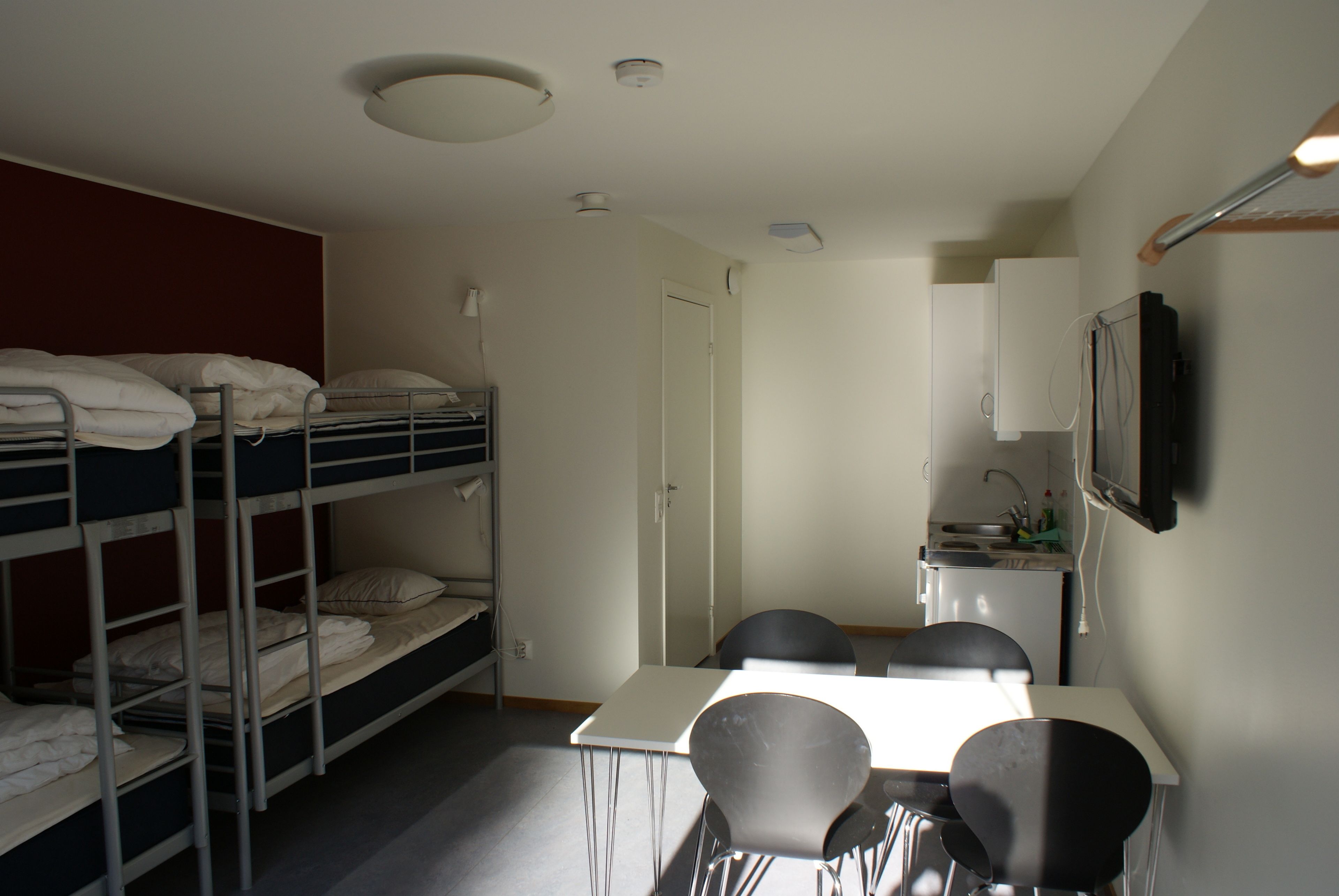 Ein Schlafsaalzimmer mit Etagenbetten, einer kleinen Küchenzeile, einem Esstisch mit Stühlen und einem an der Wand montierten Fernseher.
