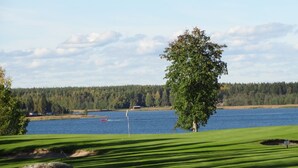 A golf course with a large tree in the foreground, a body of water, and a forested hill in the background. - Piteå Golfhotell (Pitea)