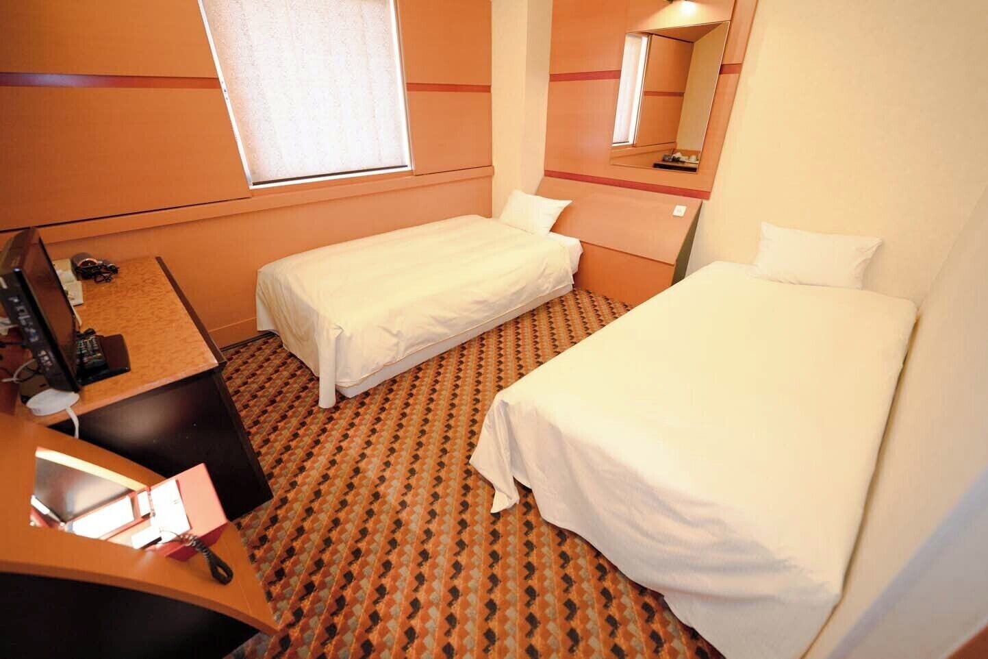 A hotel room with two single beds, a desk with a computer, a mirror, and a window.