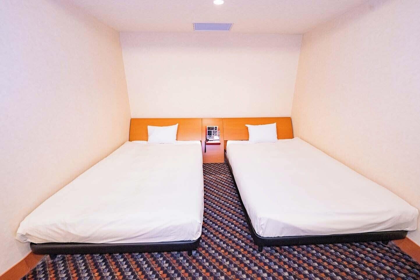 A hotel room with two single beds, a patterned carpet, and a small table with a lamp between the beds.