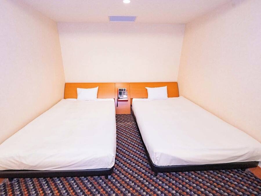 A hotel room with two single beds, a patterned carpet, and a small table with a lamp between the beds.