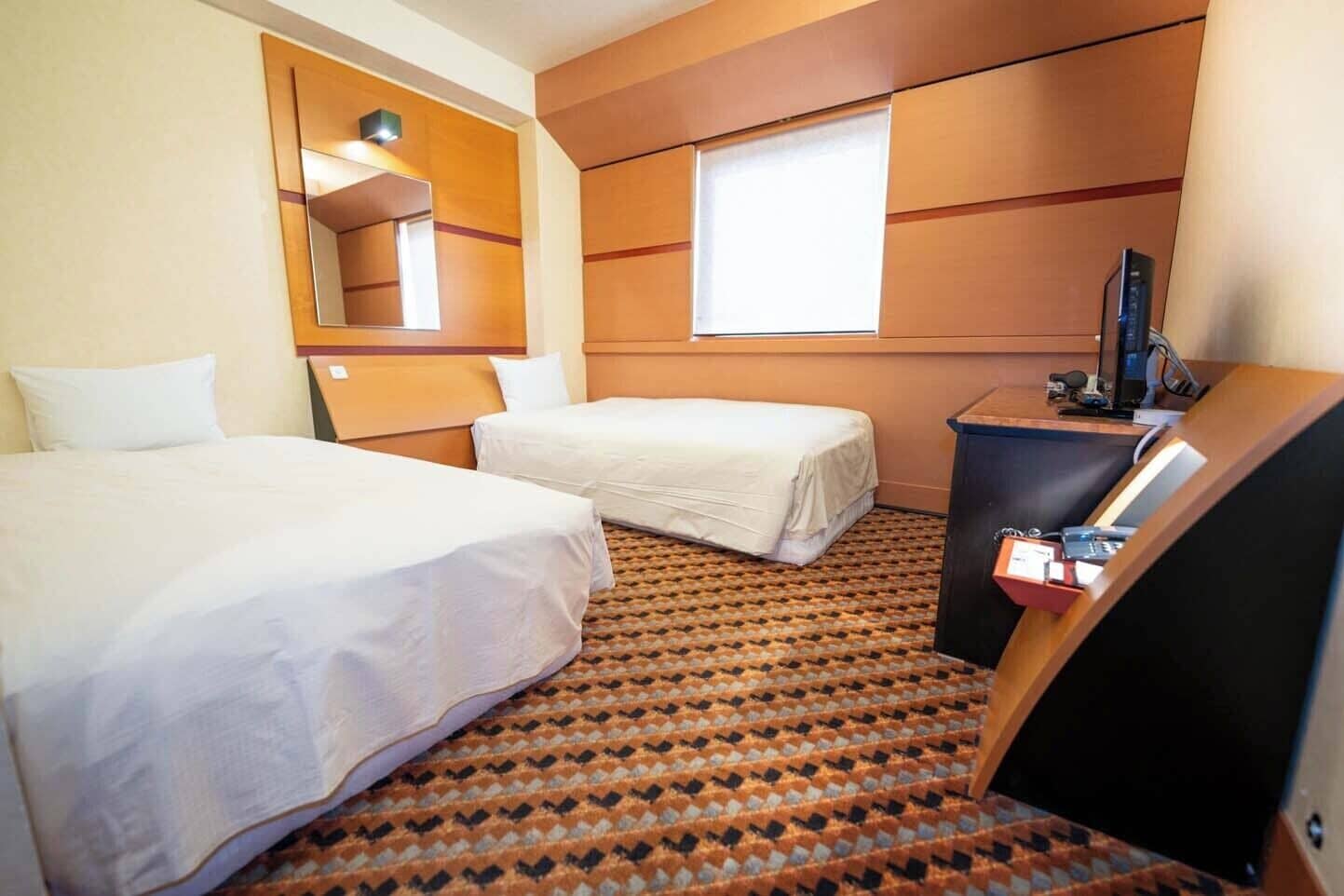 A hotel room with two beds, a television, a desk with a computer, and a window.