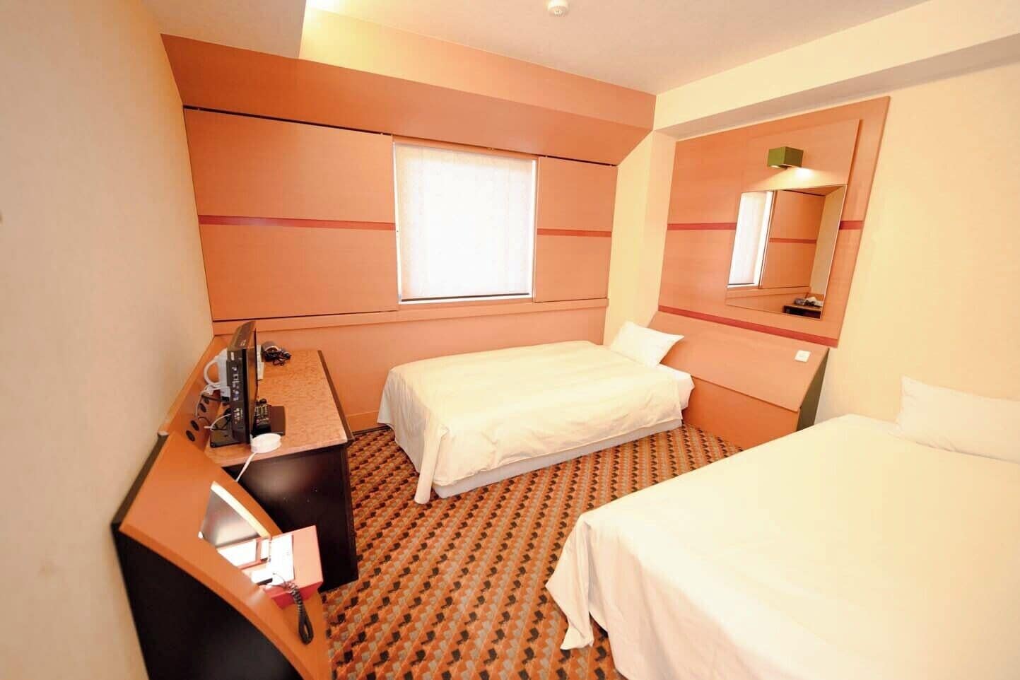 A hotel room with a desk, two beds, a window, and a mirror.
