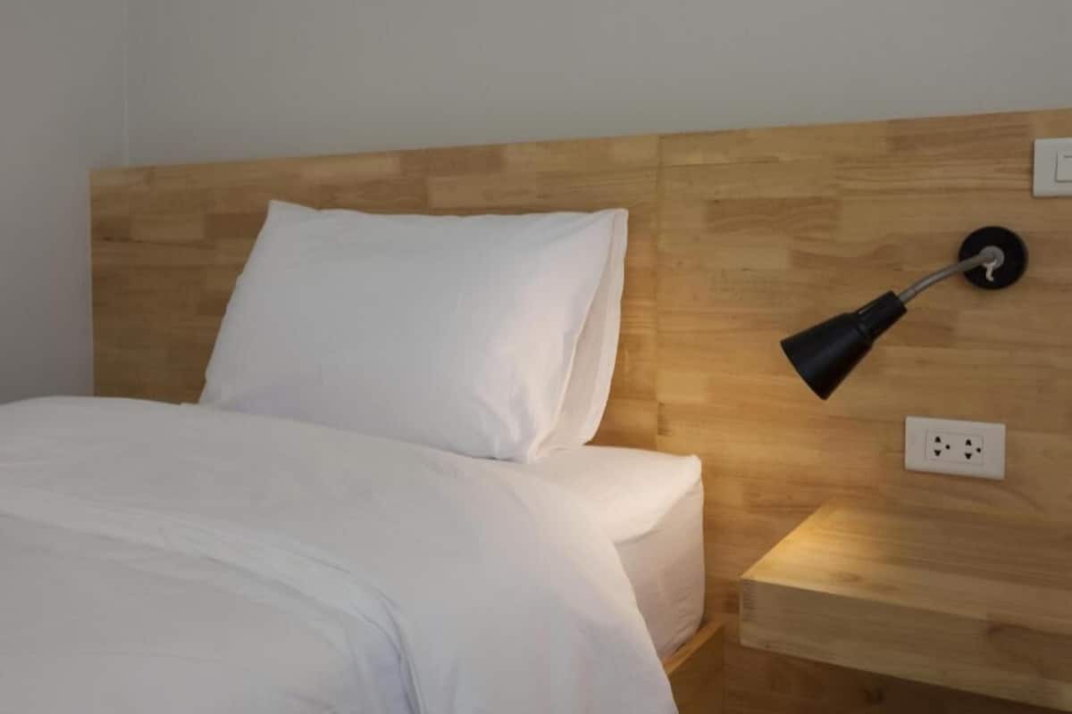 A neatly made bed with white linens, a wooden headboard, a bedside table with a lamp, and a power outlet on the wall.