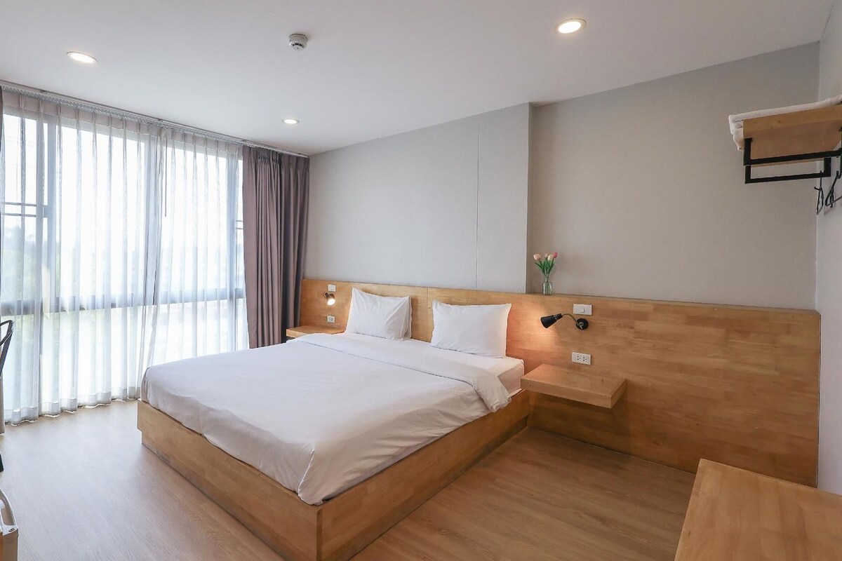 A modern hotel room with a large bed, wooden headboard, bedside tables, and a window with curtains.