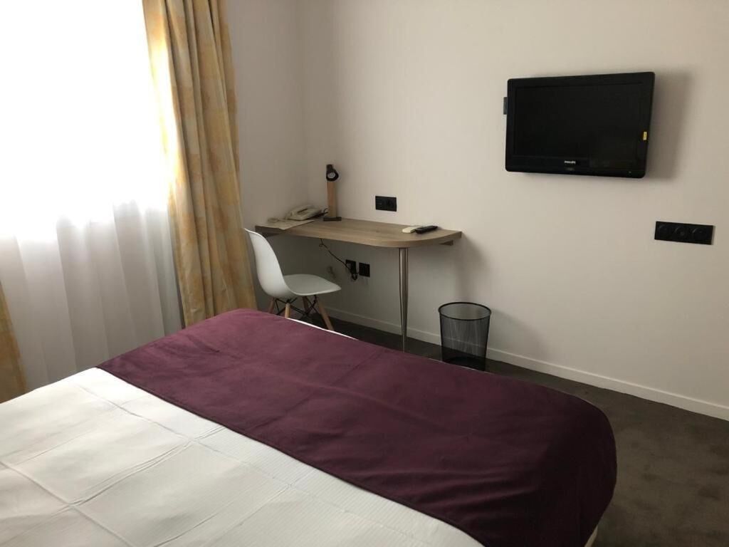 Une chambre d’hôtel avec un lit, un bureau, une chaise, une télévision et une fenêtre avec des rideaux.