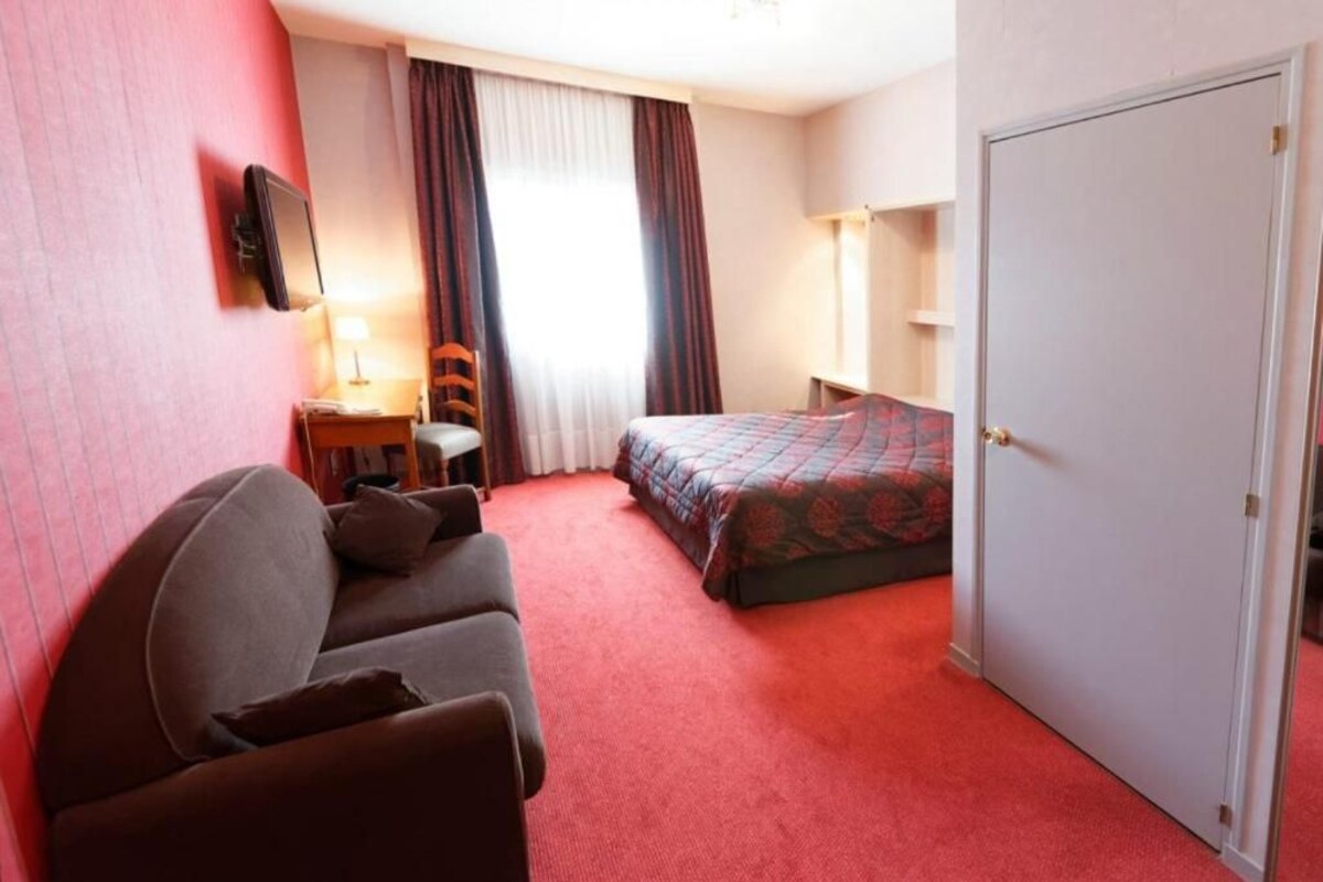 A hotel room with a bed, a sofa, a desk with a lamp, and a television on the wall.