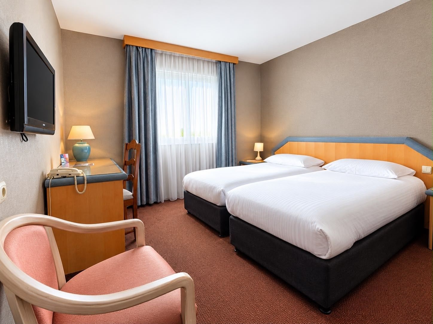 A hotel room with two beds, a desk with a telephone, a TV mounted on the wall, and a chair.