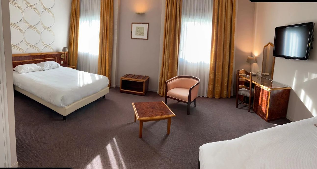 A hotel room with a bed, a desk, a chair, a small table, a television, and a mirror.