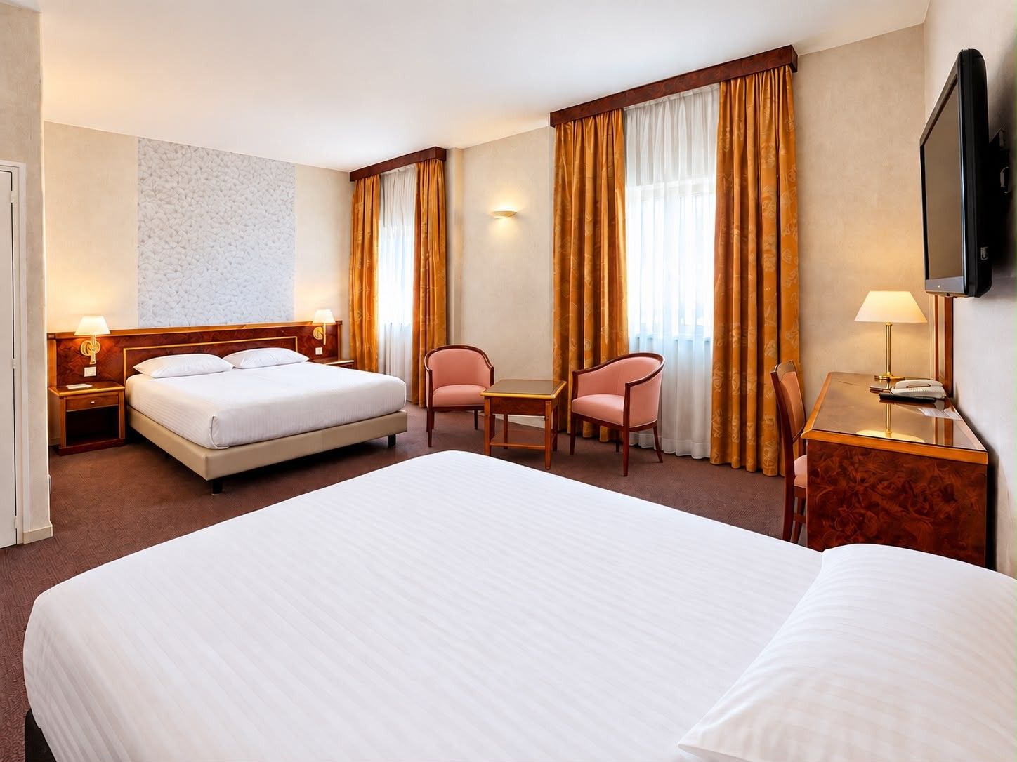 Una habitación de hotel con una cama grande, un escritorio, dos sillas, un televisor y una lámpara fijada a la pared.