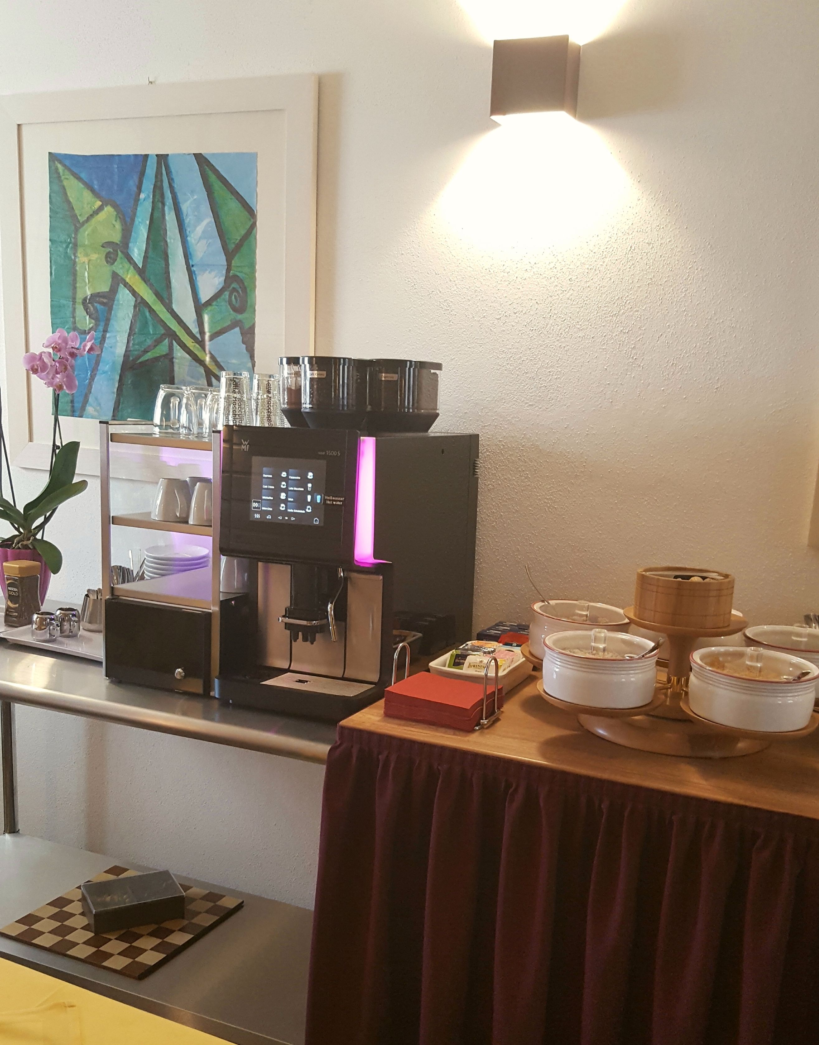 A kitchenette with a coffee machine, a small table with a red tablecloth, and a painting on the wall.