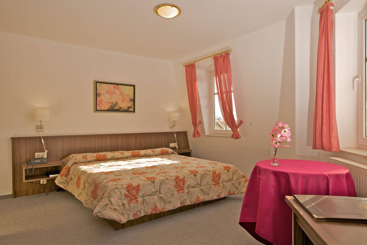 A bedroom with a bed, bedside lamps, a small table with a vase of flowers, and a window with curtains.