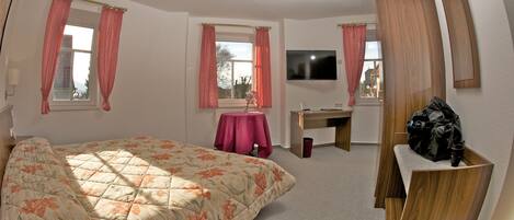 A bedroom with a bed, a desk, a chair, a TV, and a window with curtains.