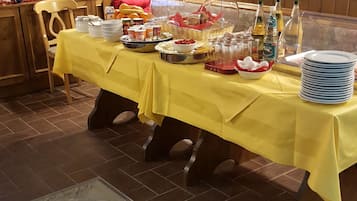 Una mesa de bufé con mantel amarillo, cubierta por una campana de cristal, y diversos alimentos y condimentos.
