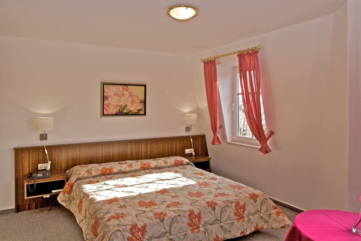 A bedroom with a wooden headboard, a floral bedspread, two bedside lamps, a nightstand, and a window with pink curtains.