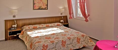 Un dormitorio con cabecera de madera, colcha estampada con motivos florales, dos lámparas de noche, una mesita de noche y una ventana con cortinas rosadas.