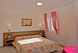 A bedroom with a wooden headboard, a floral bedspread, two bedside lamps, a nightstand, and a window with pink curtains. - Hotel Weberhof (Zittau)