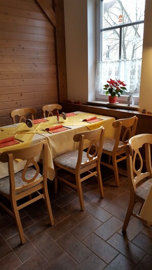 A dining area with a table set for four, wooden chairs, a window with a view of bare trees, and a potted plant on the windowsill. - Hotel Weberhof (Zittau)