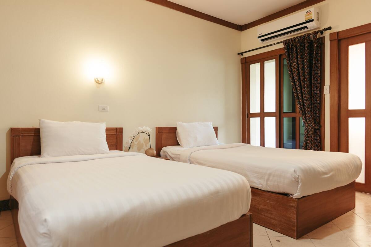 A hotel room with two single beds, a wall-mounted air conditioner, and a window with curtains.