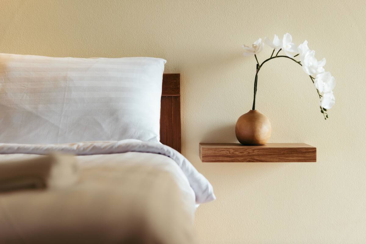 A bed with a white comforter and a wooden nightstand with a vase of white flowers.