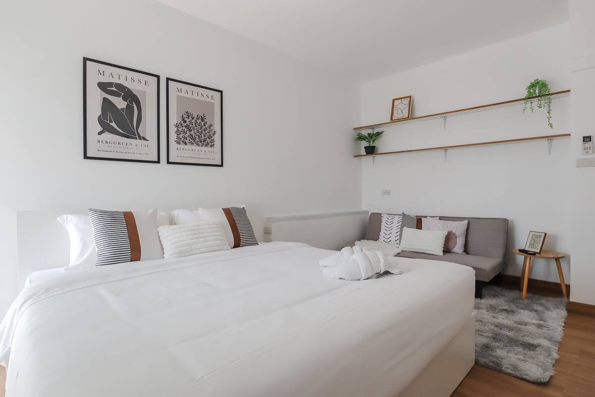 A modern bedroom with a large bed, a grey sofa, and two wall-mounted shelves with plants.
