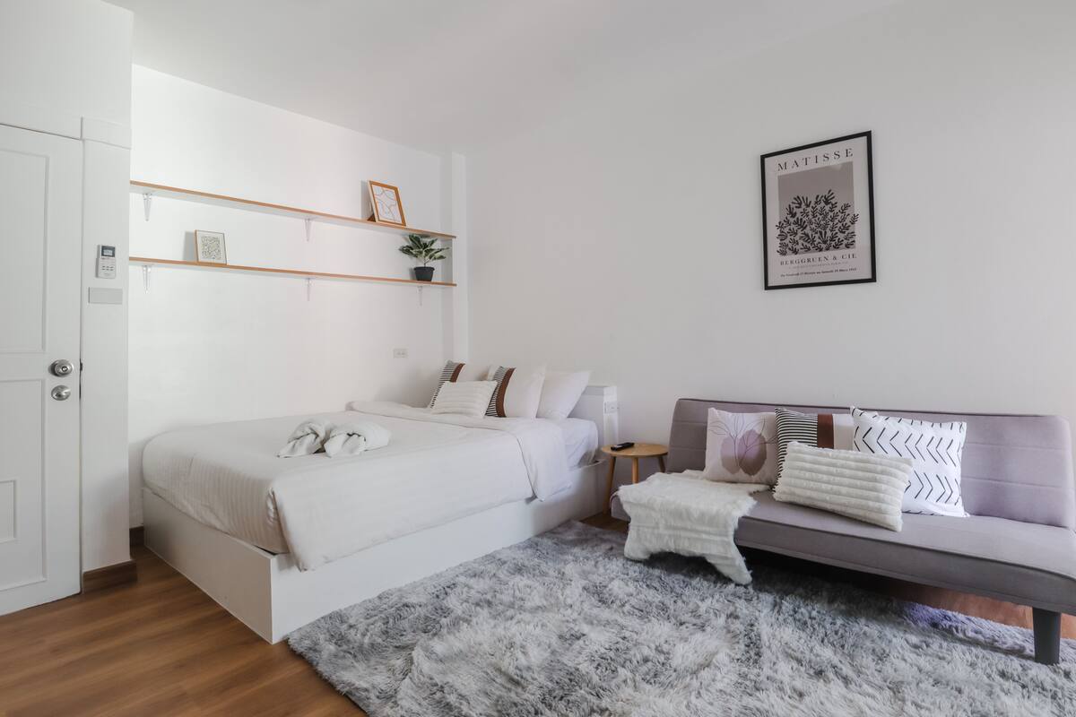 A modern bedroom with a bed, bedside table, and a sofa. There are shelves above the bed and a framed picture on the wall.