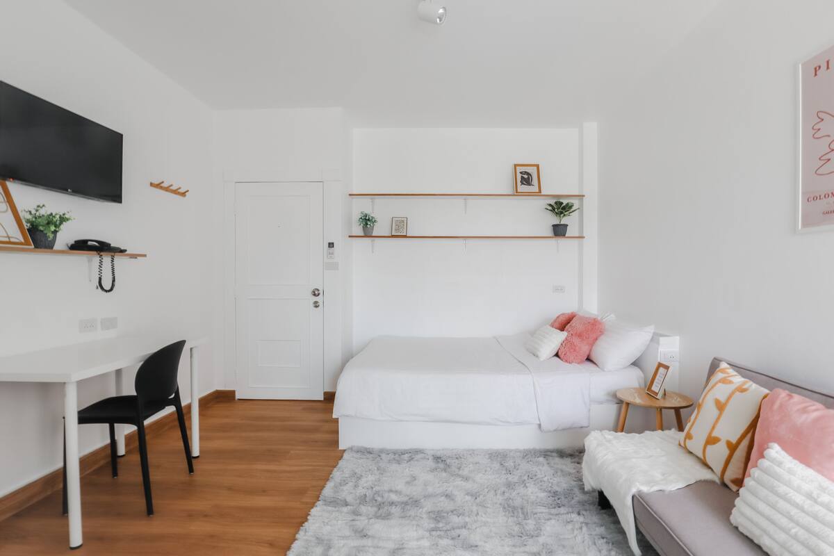 A modern bedroom with a bed, a desk, and a chair. There are shelves above the bed and a small table beside it.