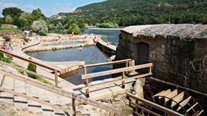 A stone building with a rustic roof, a wooden staircase leading up to a viewing platform, and a river with people in the background. - Hotel Irixo (Ourense)