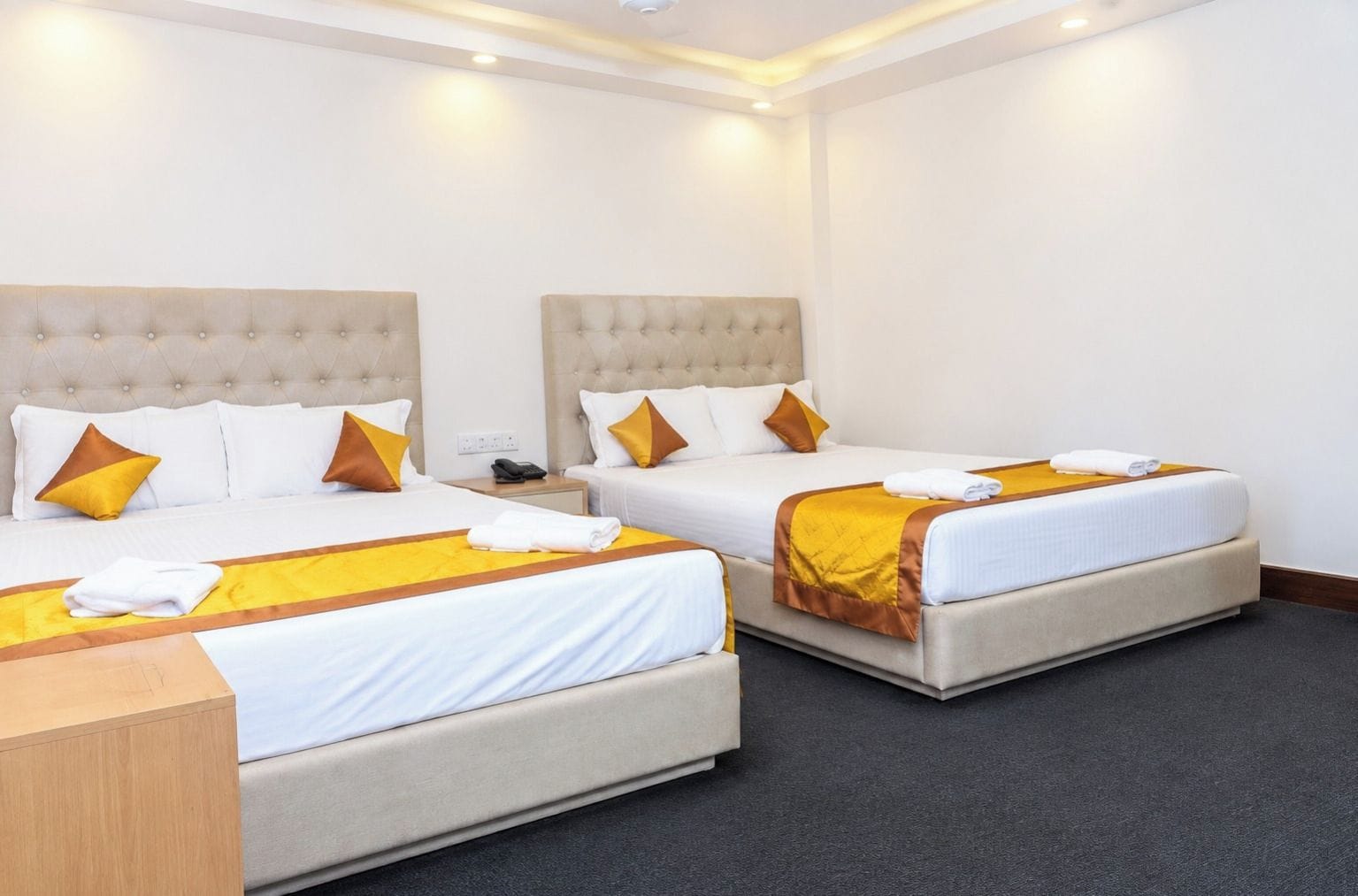 A hotel room with two beds, each with a headboard, white bedding, and brown pillows. There is a wooden nightstand with a phone and a folded towel.