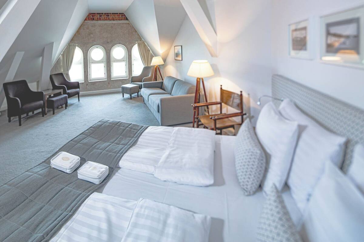 A hotel room with a large bed, a sofa, two armchairs, a wooden chair, and a view of a brick wall through arched windows.