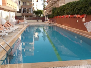A swimming pool with lounge chairs and umbrellas, surrounded by a building with balconies and a Coca-Cola banner. - Maren Beach Apart (Alanya)