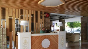A reception desk with a wooden finish and a clock above it. There are two lamps on either side of the desk, and a man is seated behind it. - Maren Beach Apart (Alanya)