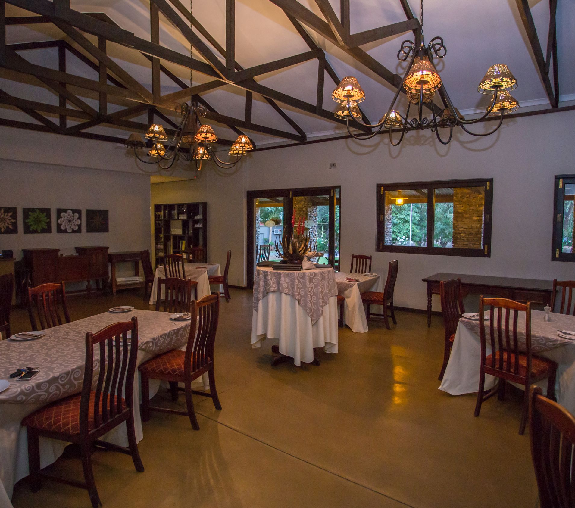 A dining area with wooden tables and chairs, a chandelier, and windows with views of greenery.