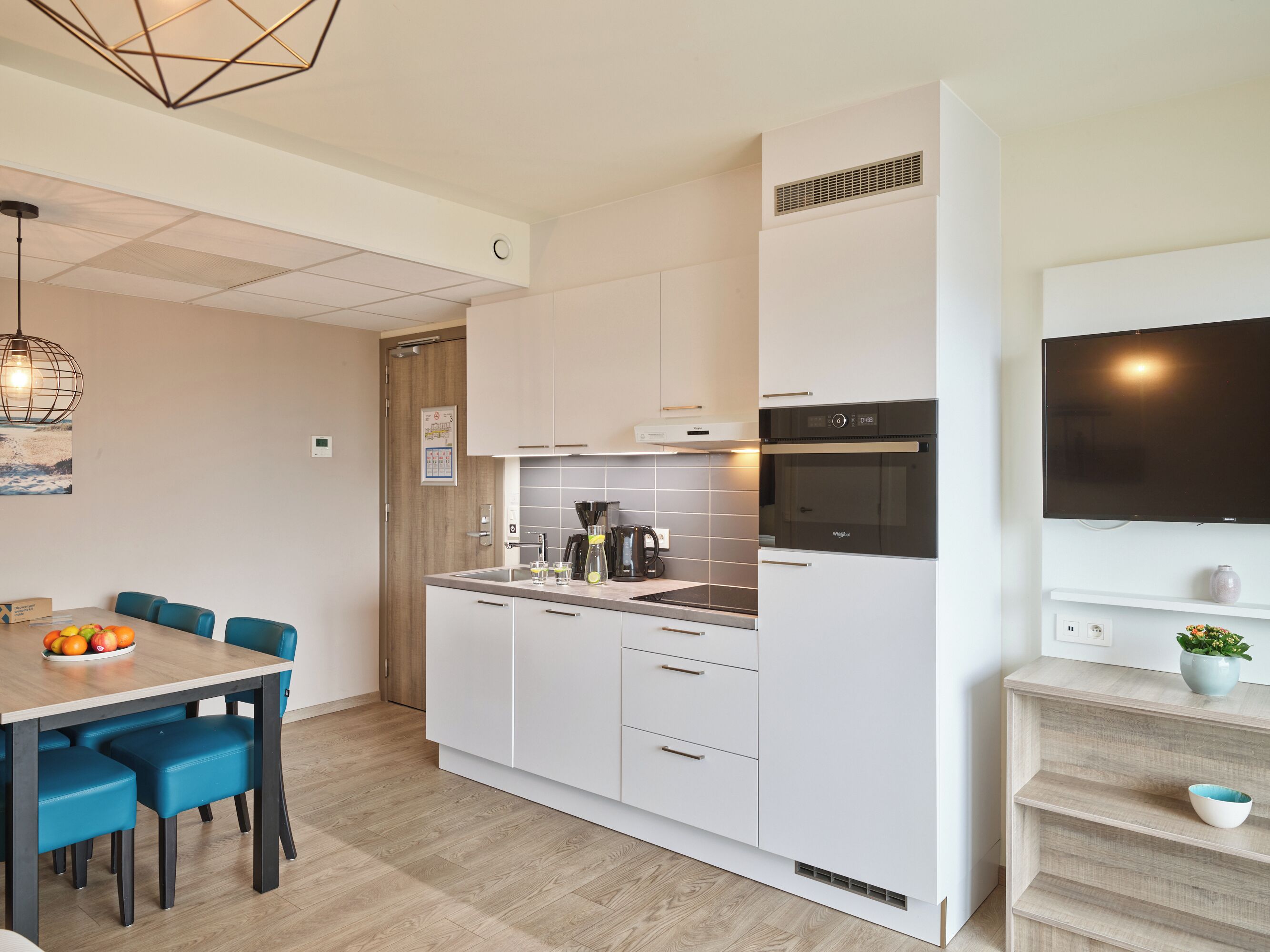 Een moderne keuken met witte kastjes, een inbouwoven en een eethoek met een houten tafel en blauwe stoelen.