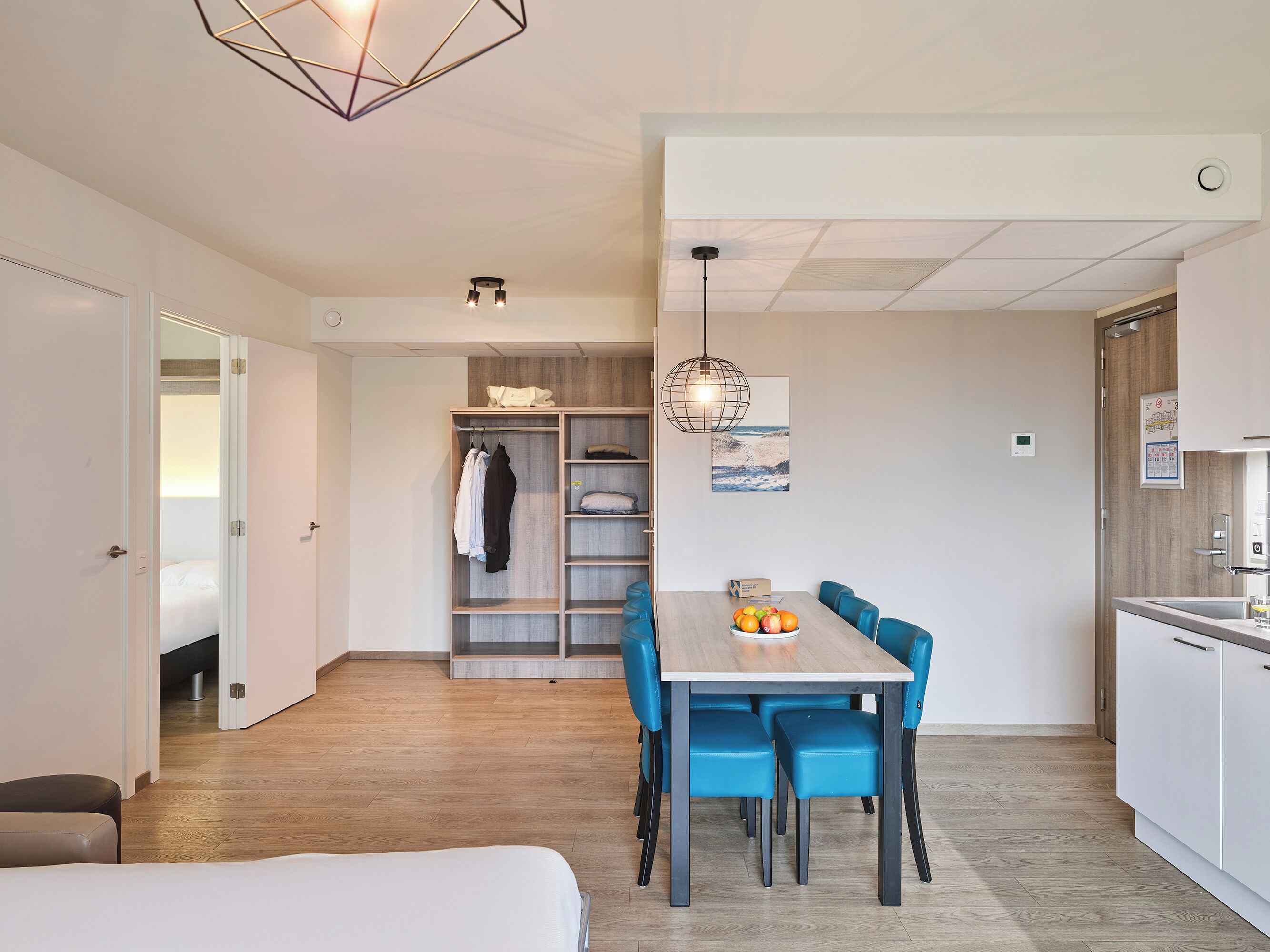 A modern hotel room with a wooden floor, a dining area with blue chairs, a white table, and a kitchen area with white cabinets.