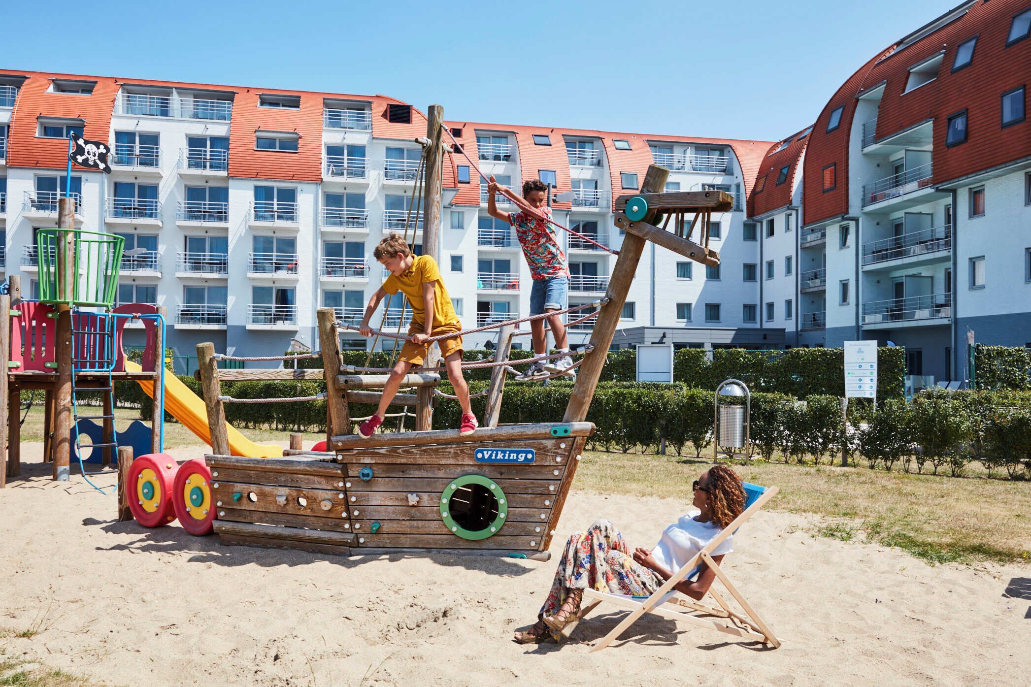 Una estructura de juego de madera con forma de barco, que incluye un letrero vikingo, toboganes y una zona de playa con una silla y una persona relajándose.