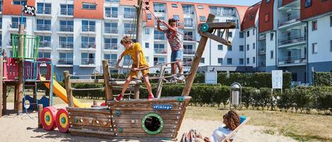 A wooden play structure resembling a ship with a Viking sign, a playground with slides, and a beach area with a chair and a person relaxing.