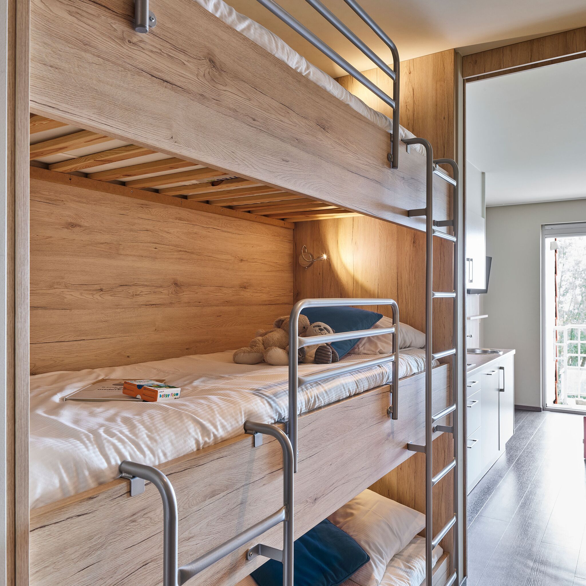 A bunk bed room with wooden walls and a kitchen area visible through an open door.