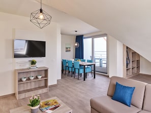 A modern living room with a flat-screen TV, a sofa, a coffee table with magazines, and a dining area with blue chairs and a table. - Holiday Suites Zeebrugge (Bruges)