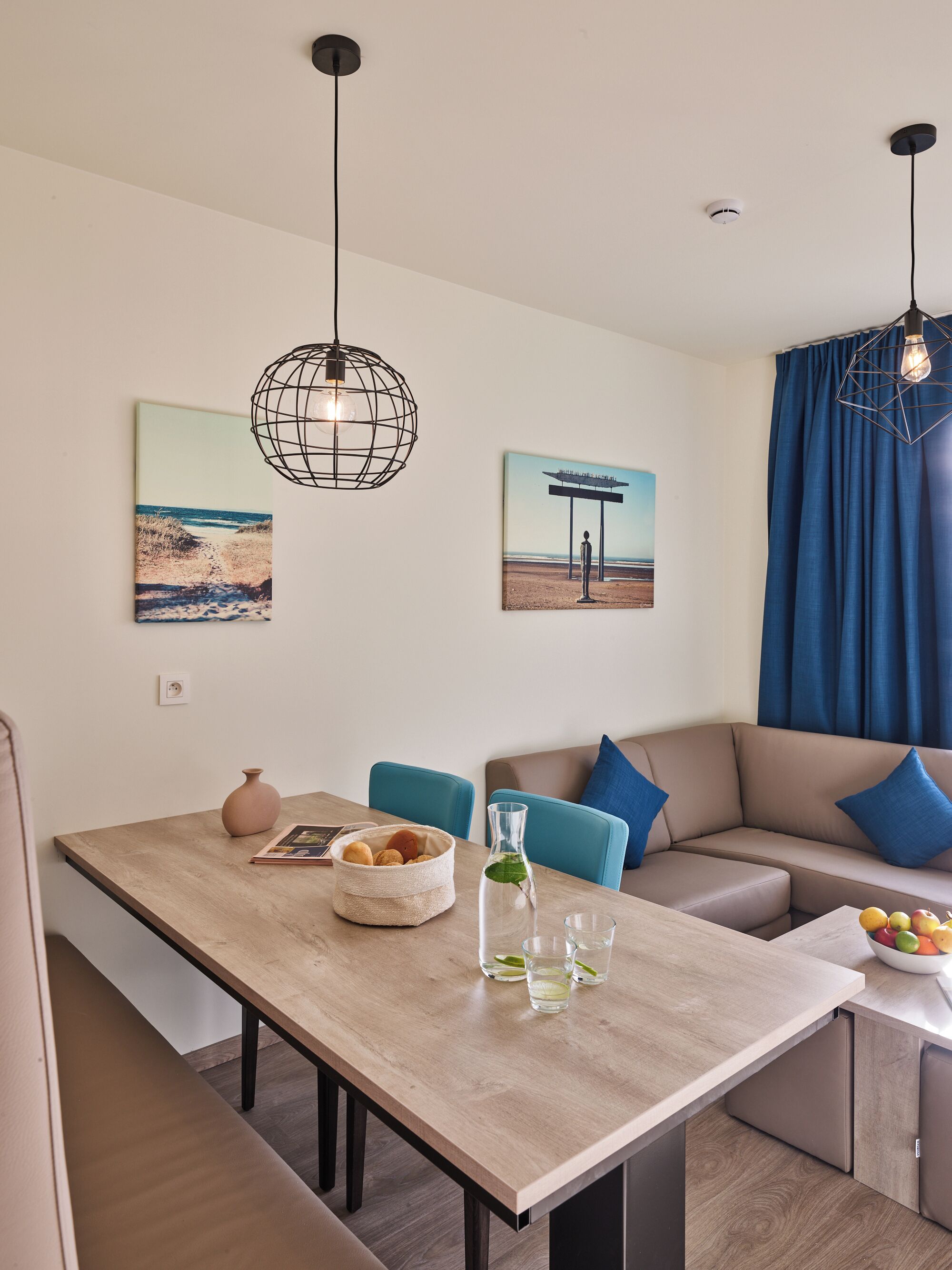 A modern living room with a wooden dining table, blue chairs, a beige sofa with blue cushions, and artwork on the walls.