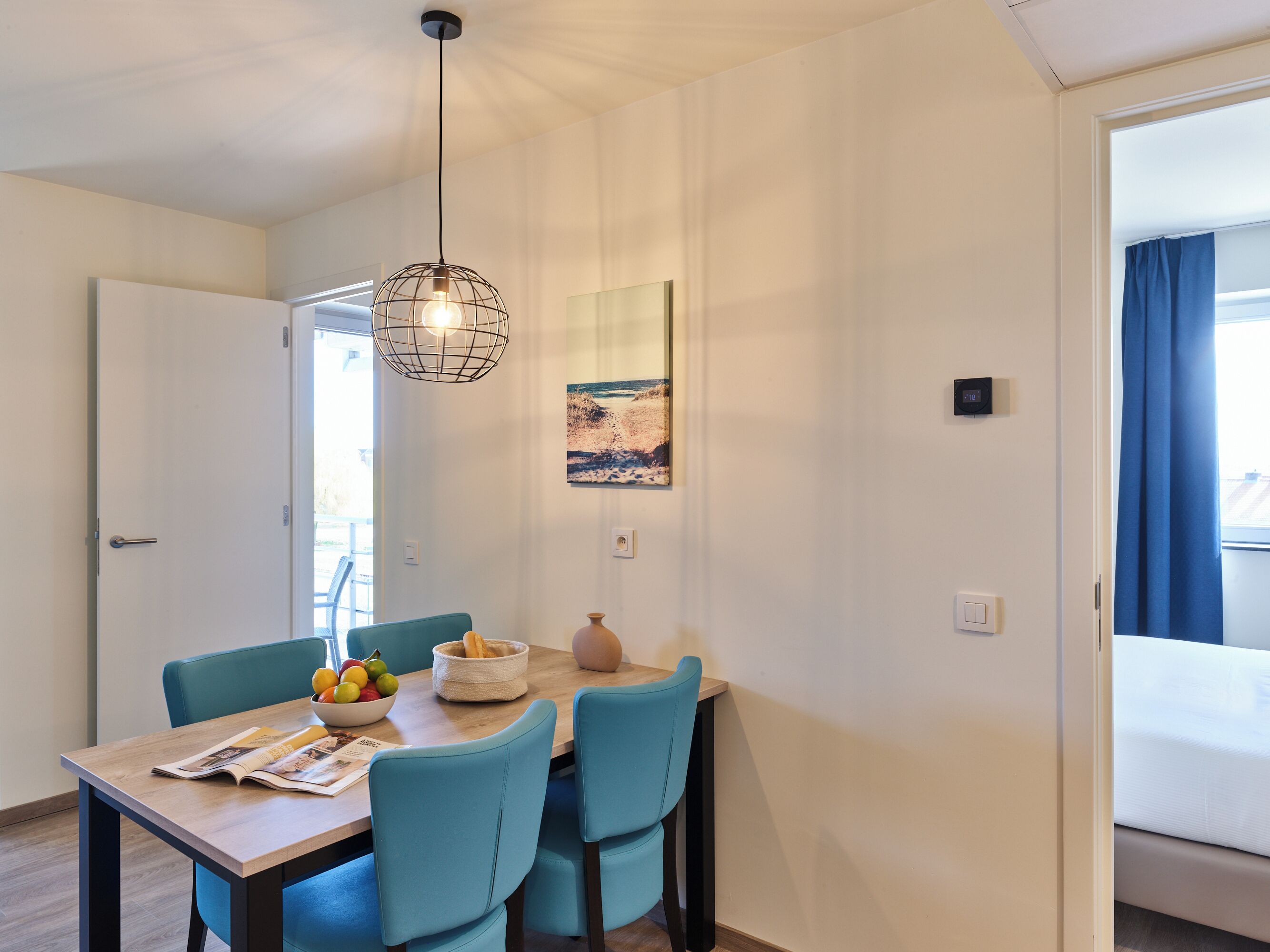 Un coin repas avec une table en bois, des chaises bleues, un saladier de fruits et un luminaire suspendu.