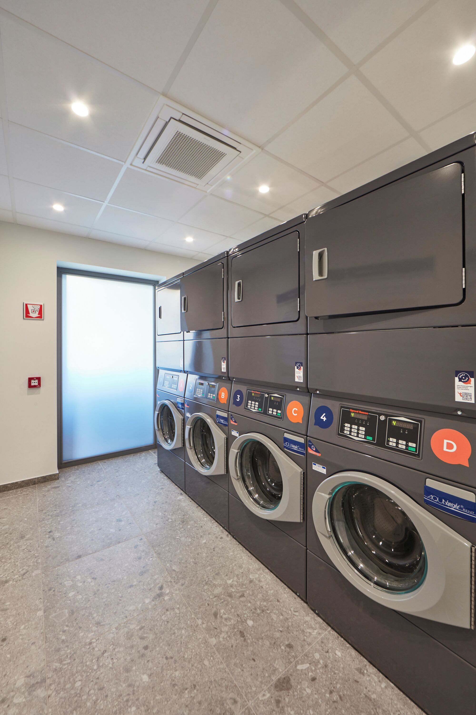 Une buanderie équipée de plusieurs machines à laver et de plusieurs sèche-linge, dotée d’un sol carrelé et d’une porte donnant sur un autre espace.