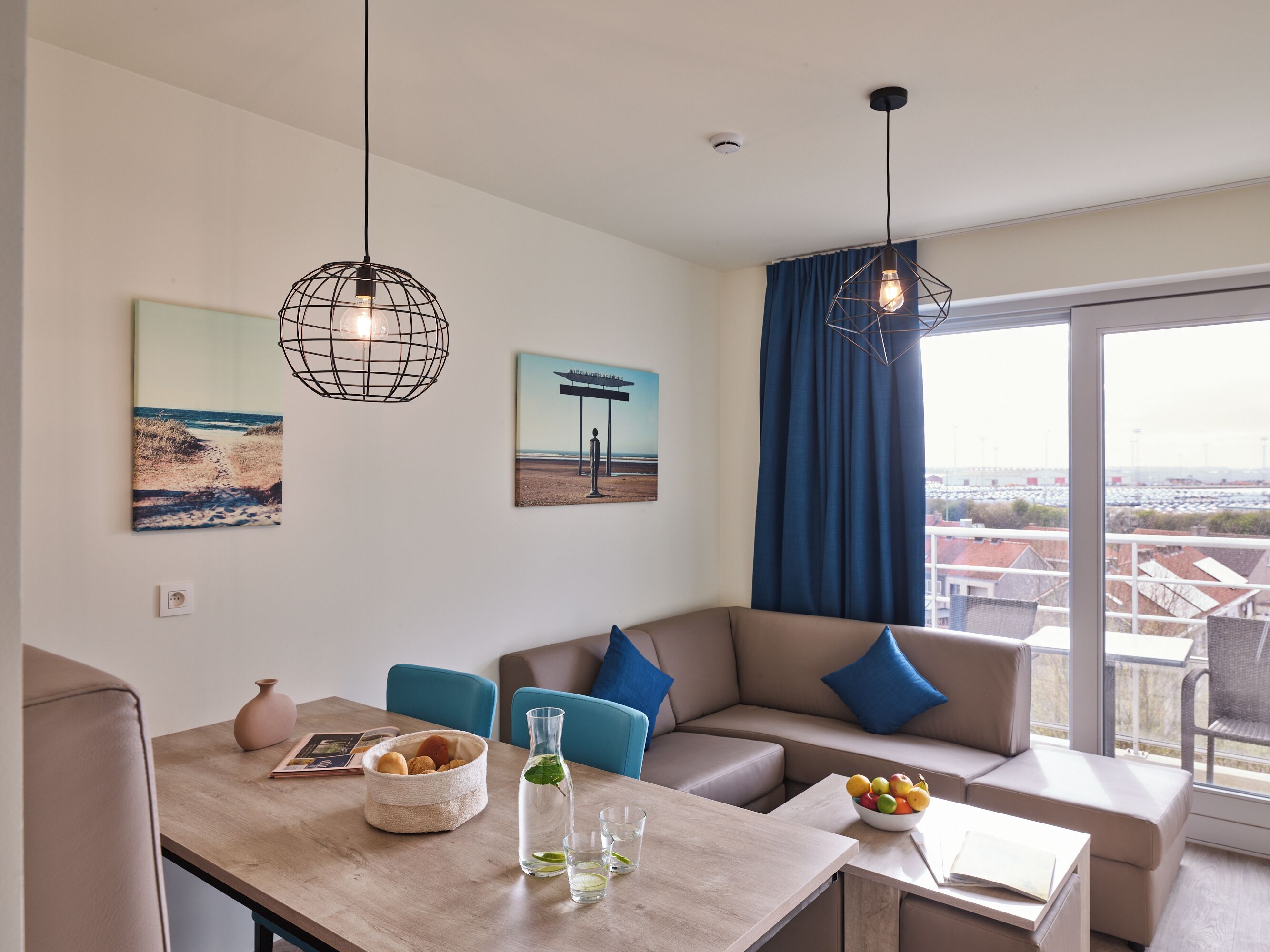 A modern living room with a wooden table, a sofa, and a large window with a view of buildings and a blue sky.