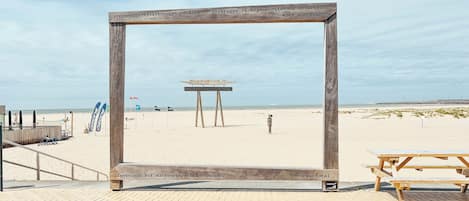 Un marco de madera con texto y hashtags en una playa, un banco de madera y un puesto de socorristas al fondo.