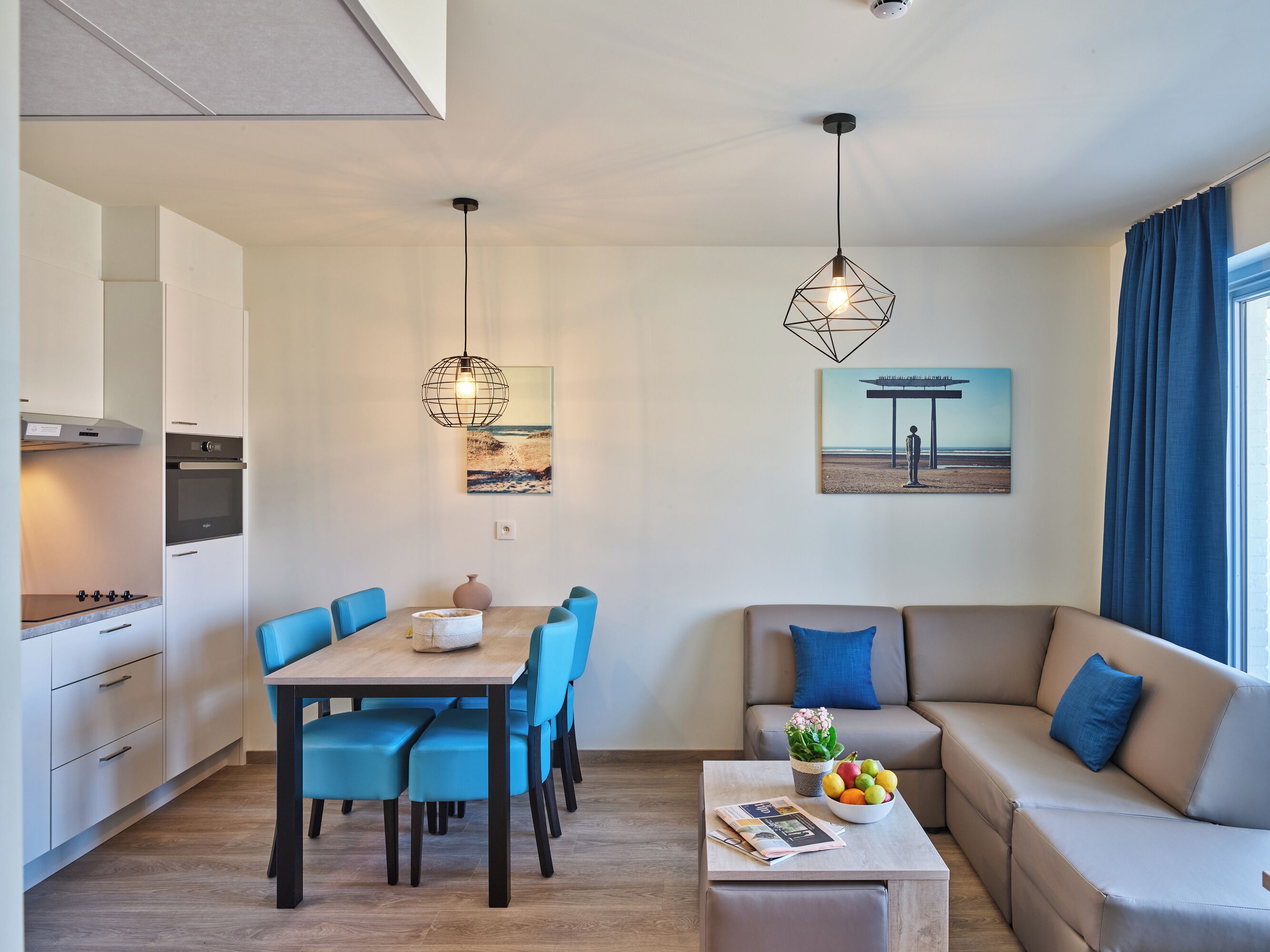 Une cuisine moderne dotée d’appareils électroménagers intégrés, un coin repas avec des chaises bleues, un canapé agrémenté d’un saladier de fruits et un tableau représentant une scène de plage.