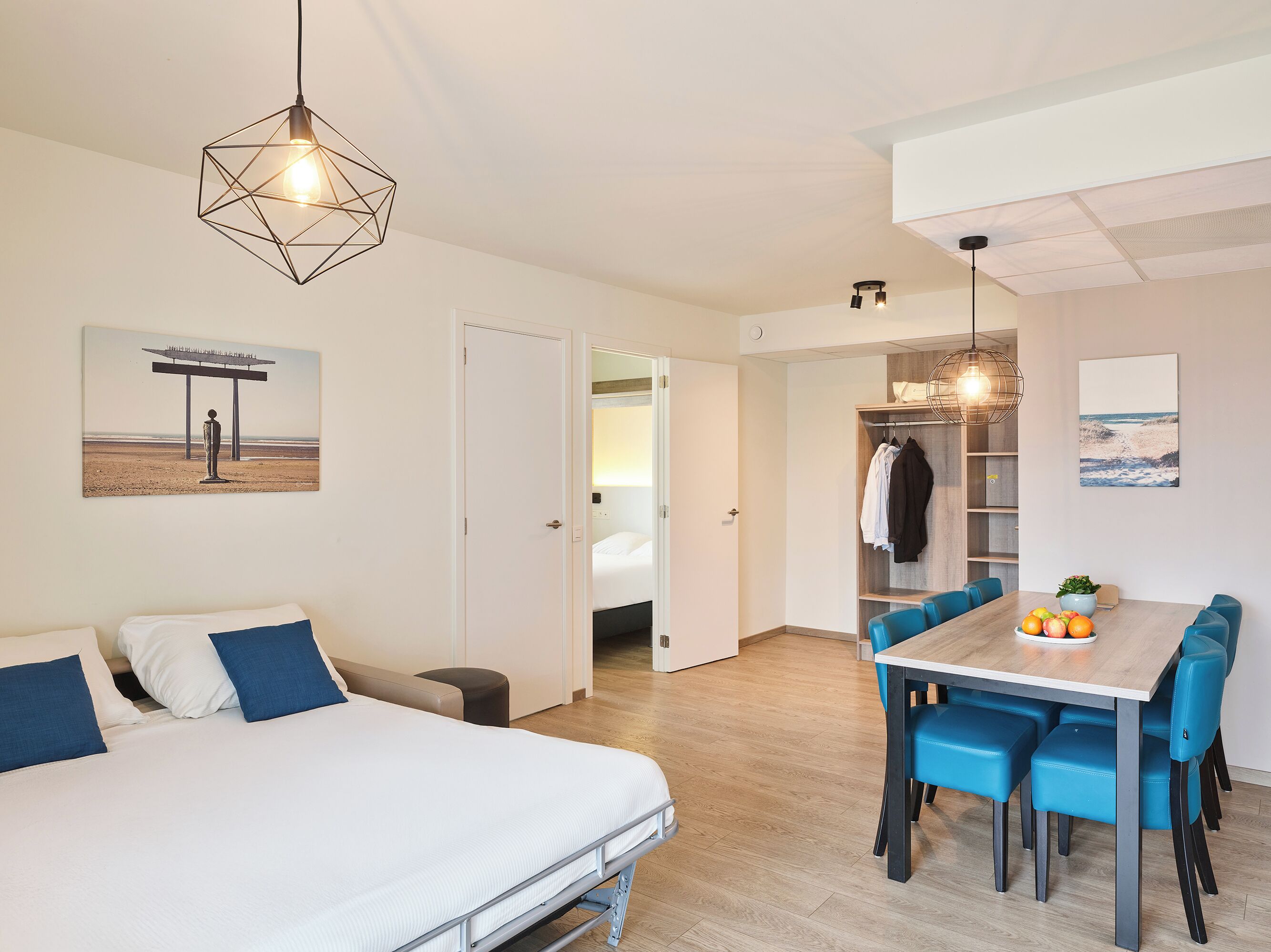 Una habitación de hotel moderna con una cama, una zona de comedor con sillas azules y vistas a la playa a través de una ventana.