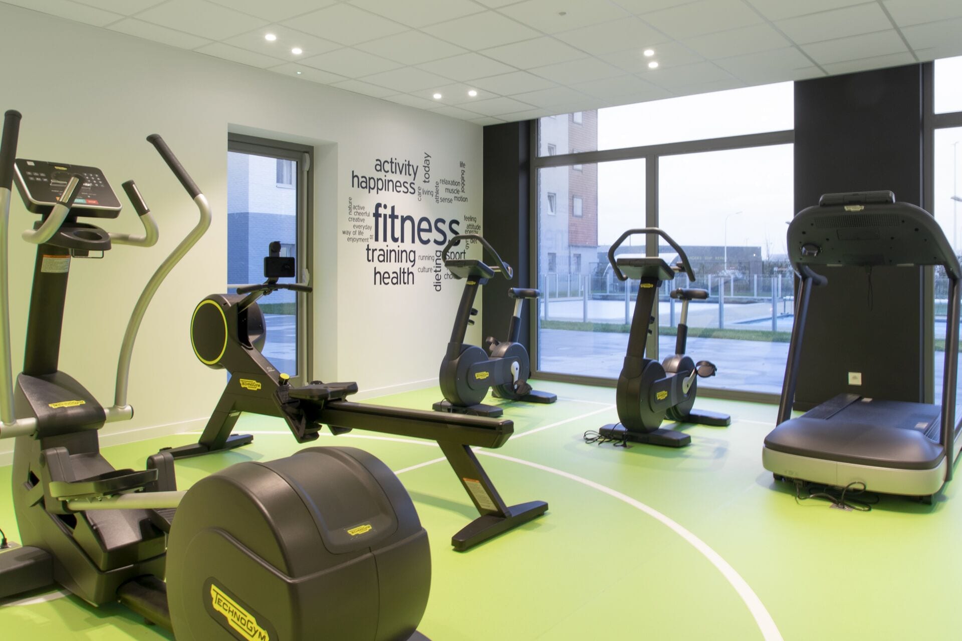 A gym with various cardio and strength training equipment, large windows, and motivational words on the wall.